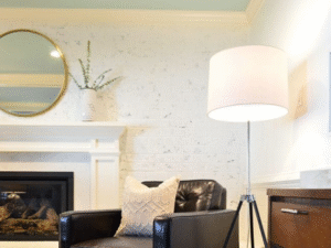 A black leather armchair with a beige pillow is beside a tripod floor lamp fitted with the Range LED Bulb 12W, placed before a fireplace and round mirror on a white brick wall.
