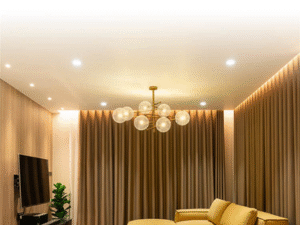 Modern living room featuring yellow sofas, a large TV to the left, a cluster chandelier, accent lighting from LED Downlight Alkor 12W, a floor plant, and beige curtains enveloping the space.
