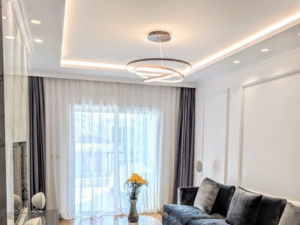 A modern living room features a grey velvet sofa, marble coffee table with yellow flowers, large window with sheer curtains, and the LED Downlight Glow 7W 3 providing warm illumination.