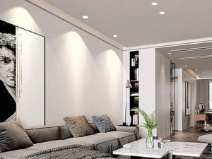 A modern living room with a gray sectional sofa, marble coffee table, wall art, and assorted cushions is brightly lit by the LED Downlight Mercury Plus 12W. Neutral tones and contemporary decor enhance the stylish, inviting space.
